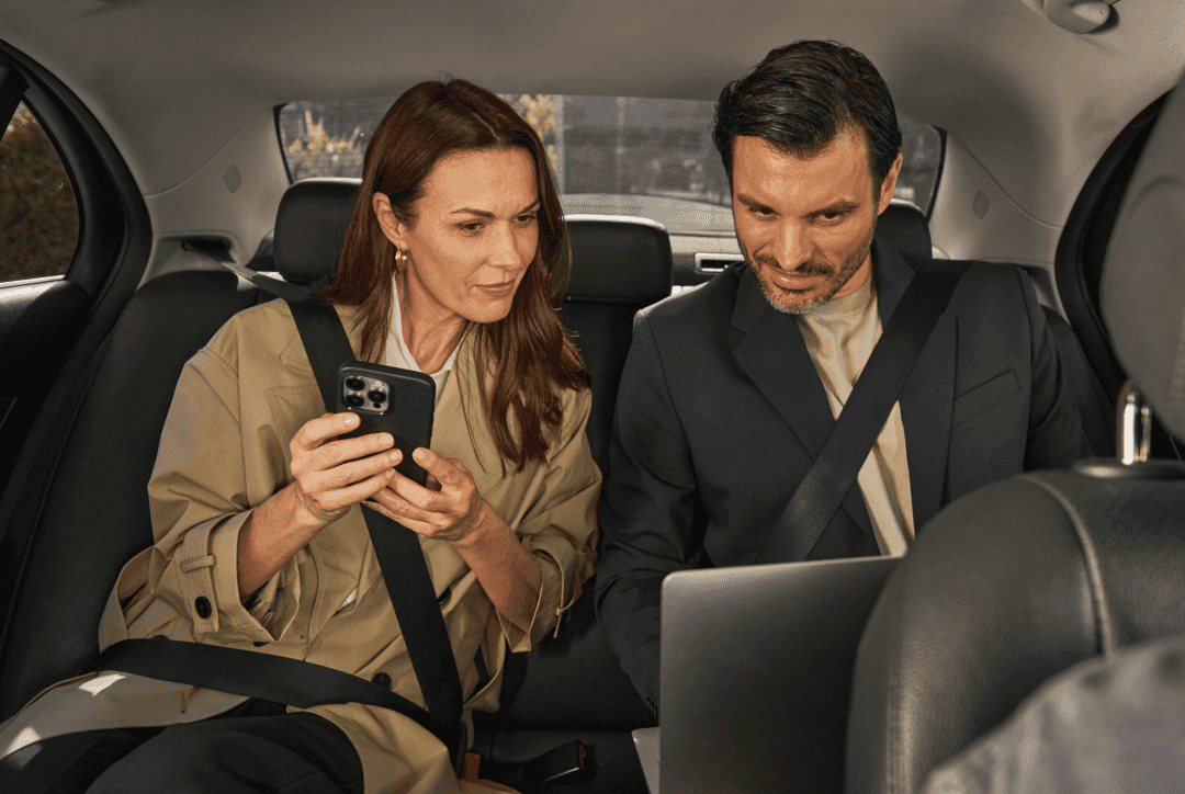 Zwei Angestellte schauen auf ein Telefon mit einem FREENOW-Logo auf dem Bildschirm und einem roten Taxi im Hintergrund